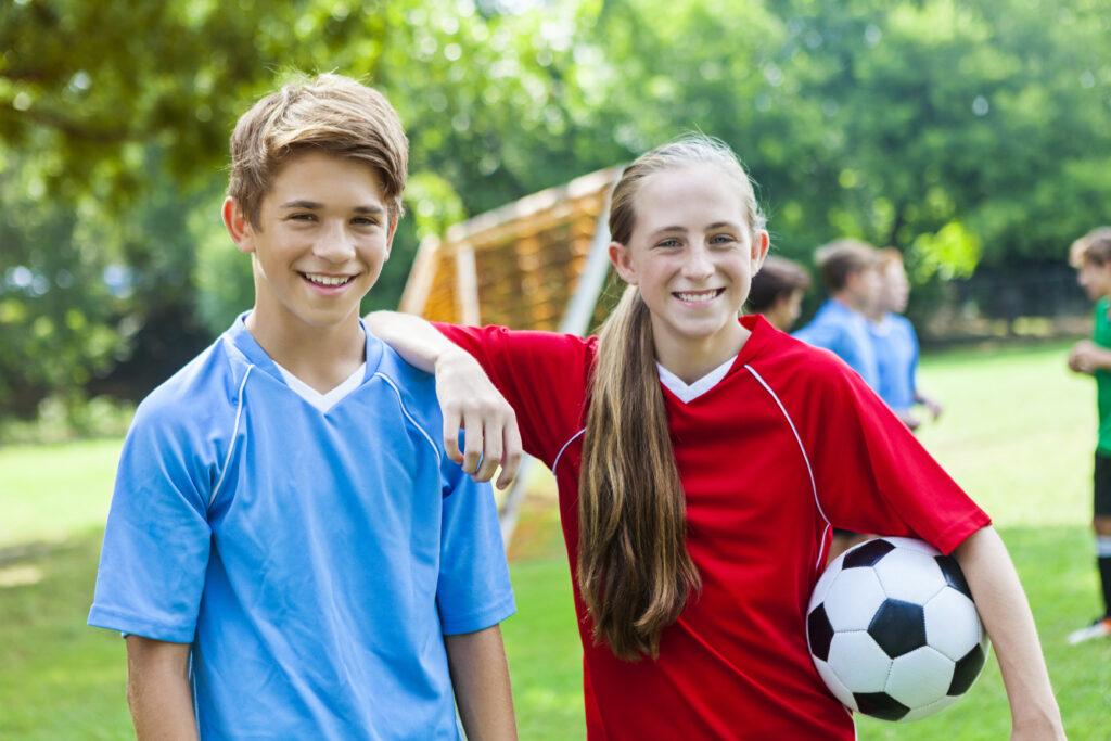 Teenage brother and sister or soccer friends kid around before soccer practice. The girl has her arm on the boy's shoulder and is holding a soccer ball. She has long blond hair in a ponytail across her shoulder. She is wearing a bright red jersey. The boy is wearing a blue jersey and has short brown hair. They are both smiling. Players are on the field in the background along with the soccer goal.