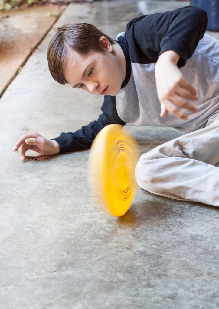 vertical orientation close up of a teenage boy with autism and down's syndrome, staring intently as he is laying on a cement floor, spinning a yellow disc, with the disc in motion, and copy space